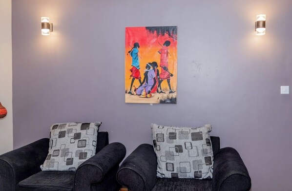 A room with two black sofas, each with a patterned pillow, and a colorful painting on the wall. - Lux Suites Enkarsis Bungalow Kisaju (Kitengela)