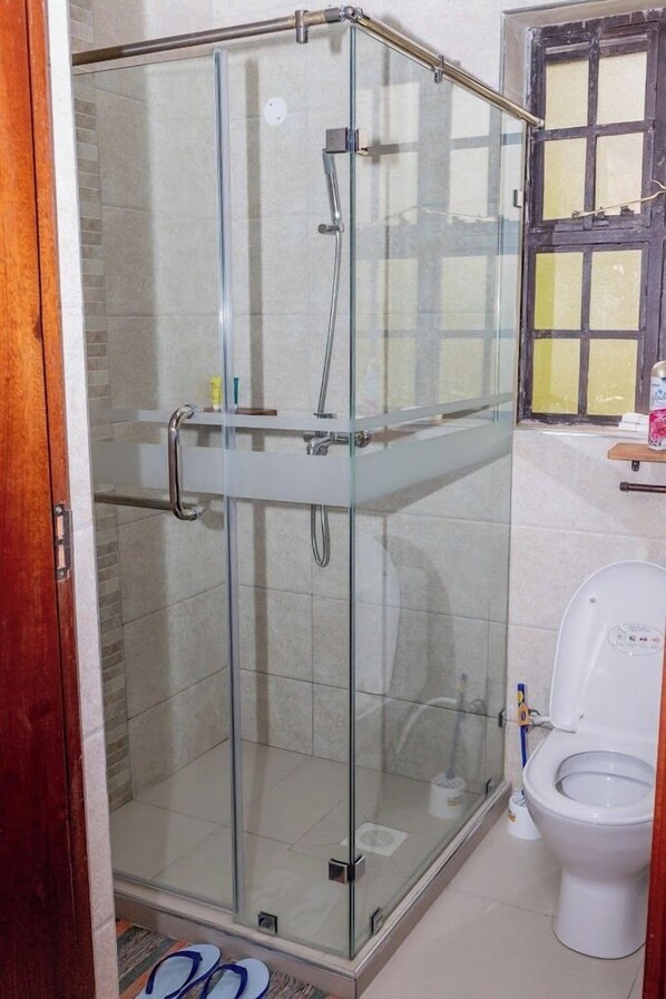 Ducha con cerramiento de vidrio, inodoro blanco y ventana con diseño de cuadrícula.