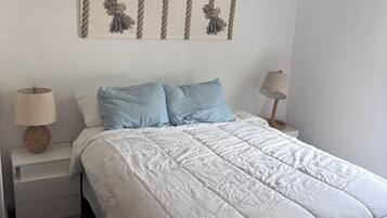 A bedroom with a white bed, two blue pillows, a ceiling fan, and a wall-mounted artwork.