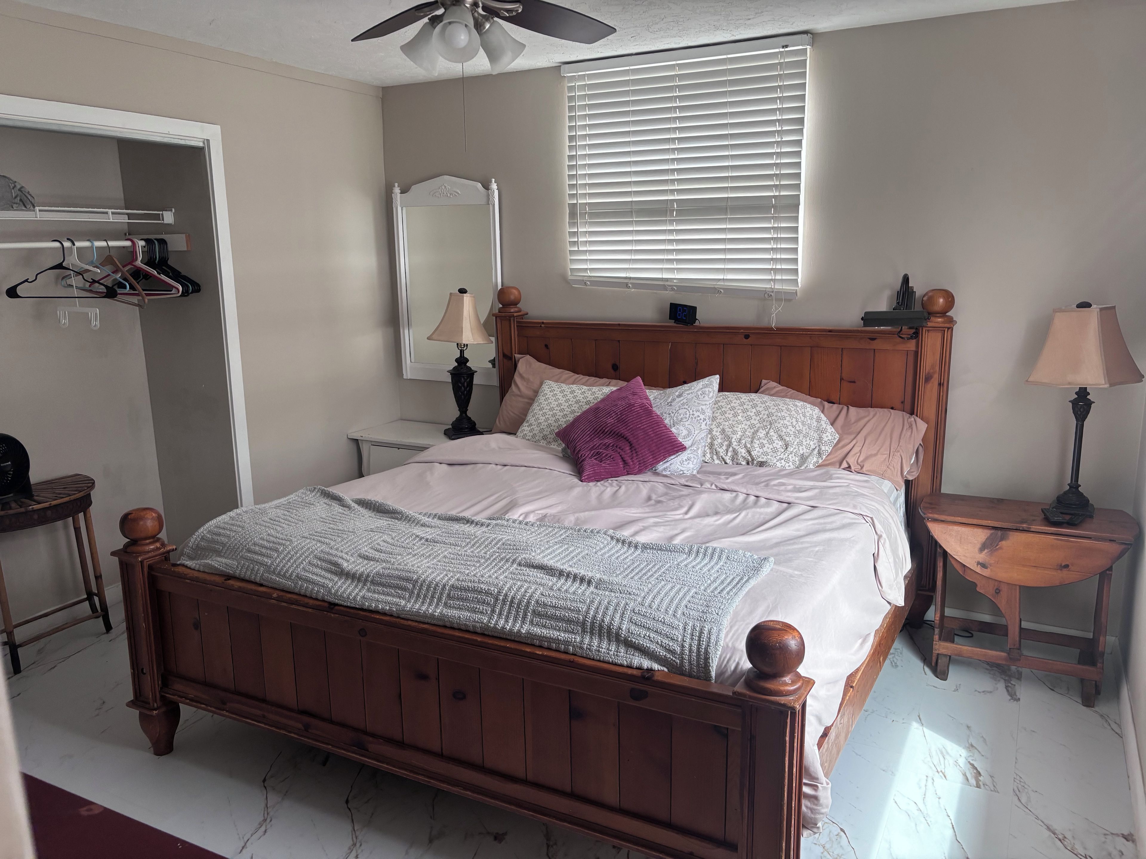 A bedroom with a wooden bed, a bedside table with a lamp, a mirror, a wardrobe, and a ceiling fan.