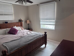 Un dormitorio con cama de madera, una mesita de noche con lámpara, ventilador de techo y ventana con persianas.