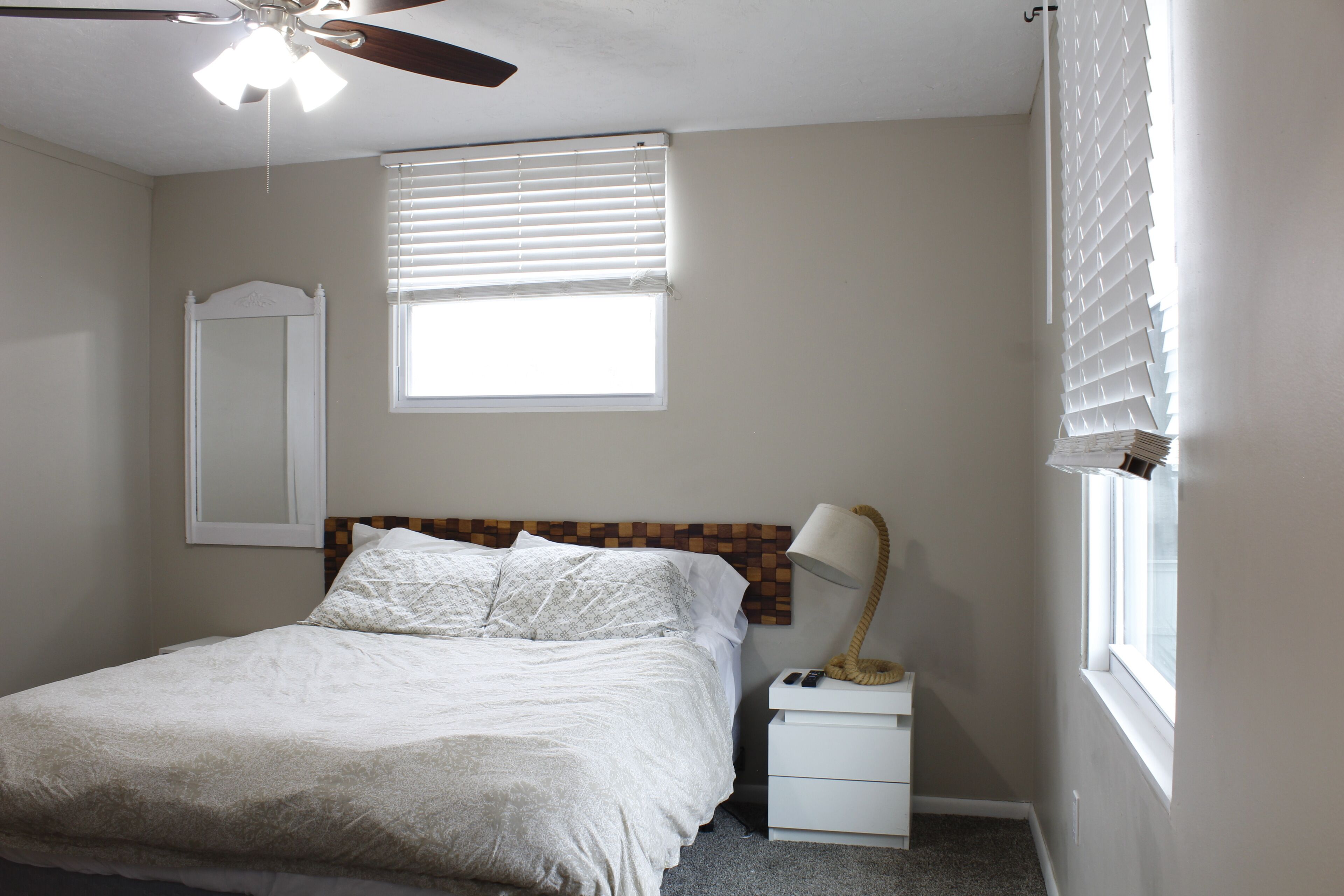 A bedroom with a bed, a nightstand, a lamp, a ceiling fan, and two windows with blinds.