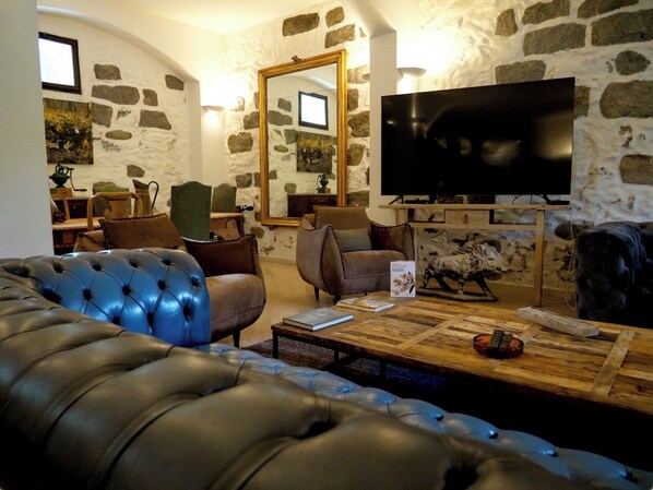 A cozy living room with a stone wall, a large flat-screen TV, leather sofas, and a coffee table with books and a remote control. - Auberge u Mulinu (Casalabriva)