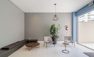 Uma área de espera moderna com um banco, duas cadeiras, uma mesinha, uma planta em vaso e um pendente.