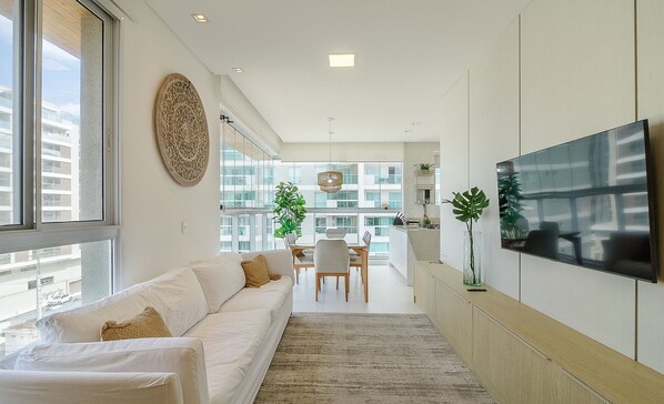 A modern living room with a sofa, a dining area, and a TV mounted on the wall. - Apartment w/ Barbecue 240m From the Beach Ehi0401 (Itajai)