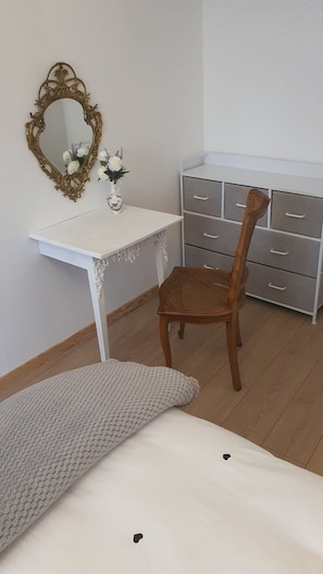 Et soveværelse med en hvid kommode, en træstol, et spejl med dekoreret ramme og en lille vase med blomster.