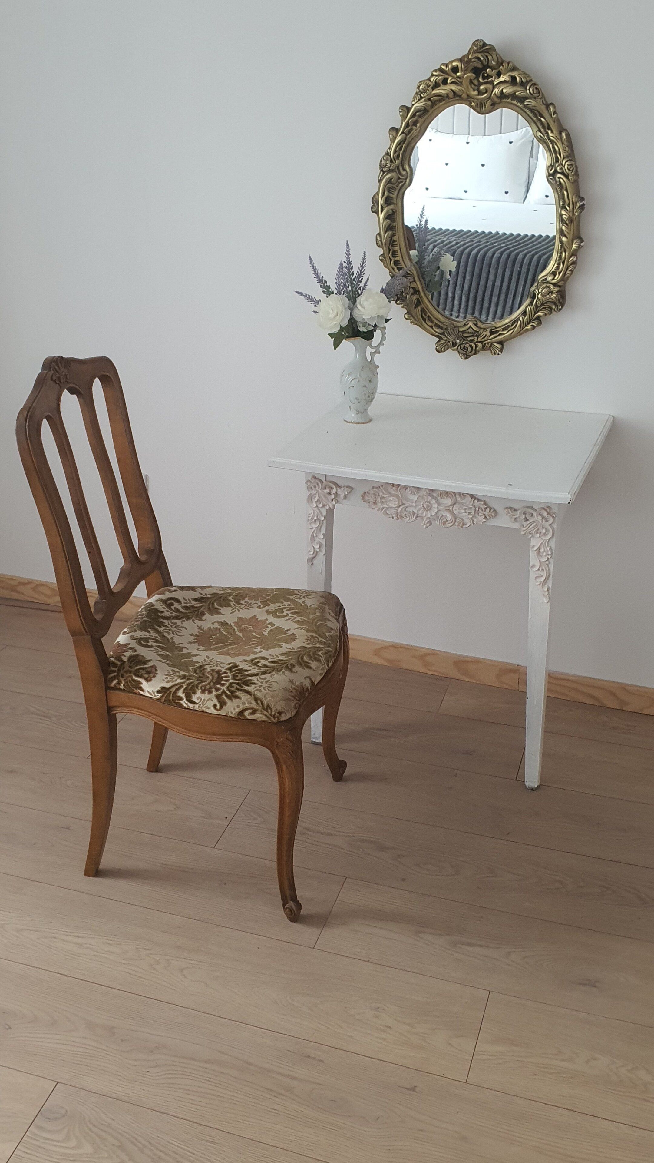 A wooden chair with a floral cushion, a white table with ornate carvings, and a gold-framed mirror on the wall.