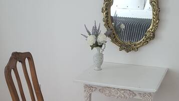 A wooden chair with a floral cushion, a white table with ornate carvings, and a gold-framed mirror on the wall.