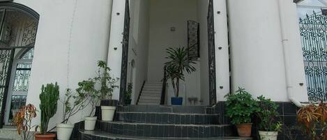 A building with a grand entrance featuring a staircase with potted plants, white columns, and a metal gate.