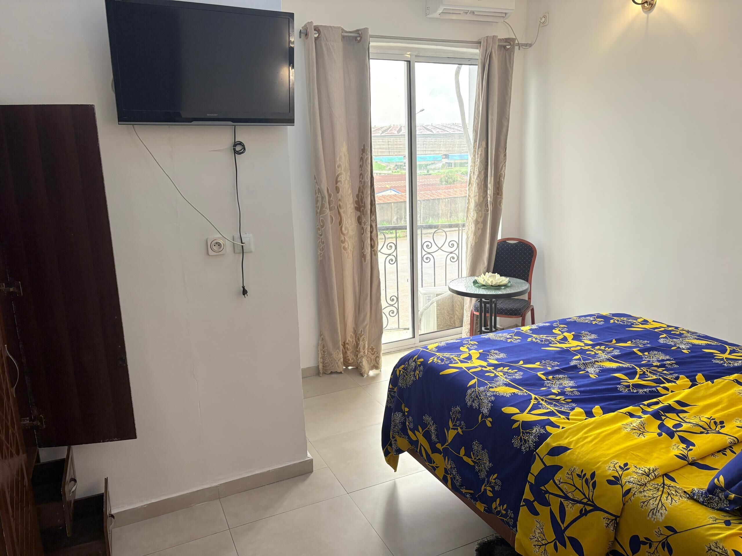 Et hotelværelse med en seng, et fjernsyn ophængt på væggen, et lille bord med en stol og en balkon med gardiner.