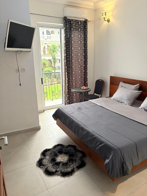 A hotel room with a bed, a TV mounted on the wall, a small table with a chair, and a view of the outside balcony. - Jacobs Hôtel (Douala)