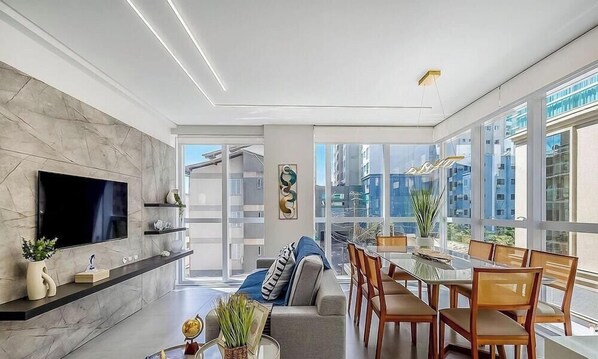 A modern living room with a flat-screen TV, a sofa, a dining area with wooden chairs, and large windows offering a city view. - Modern 3BR apt Near the Beach Ema0201 (Itapema)