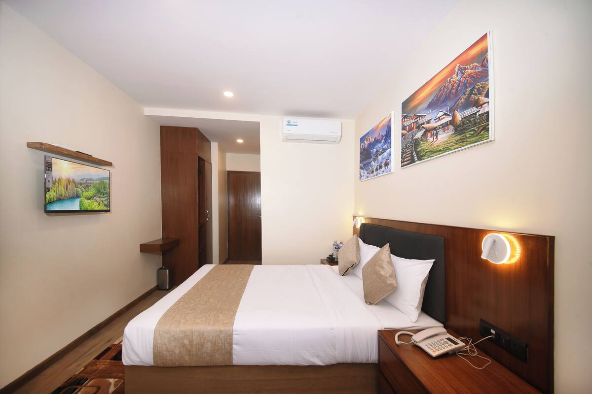 Una habitación de hotel con cama, mesita de noche, aire acondicionado de pared y dos cuadros enmarcados.