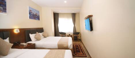 A hotel room with two beds, a desk with a lamp, a chair, a TV mounted on the wall, and a window with curtains.