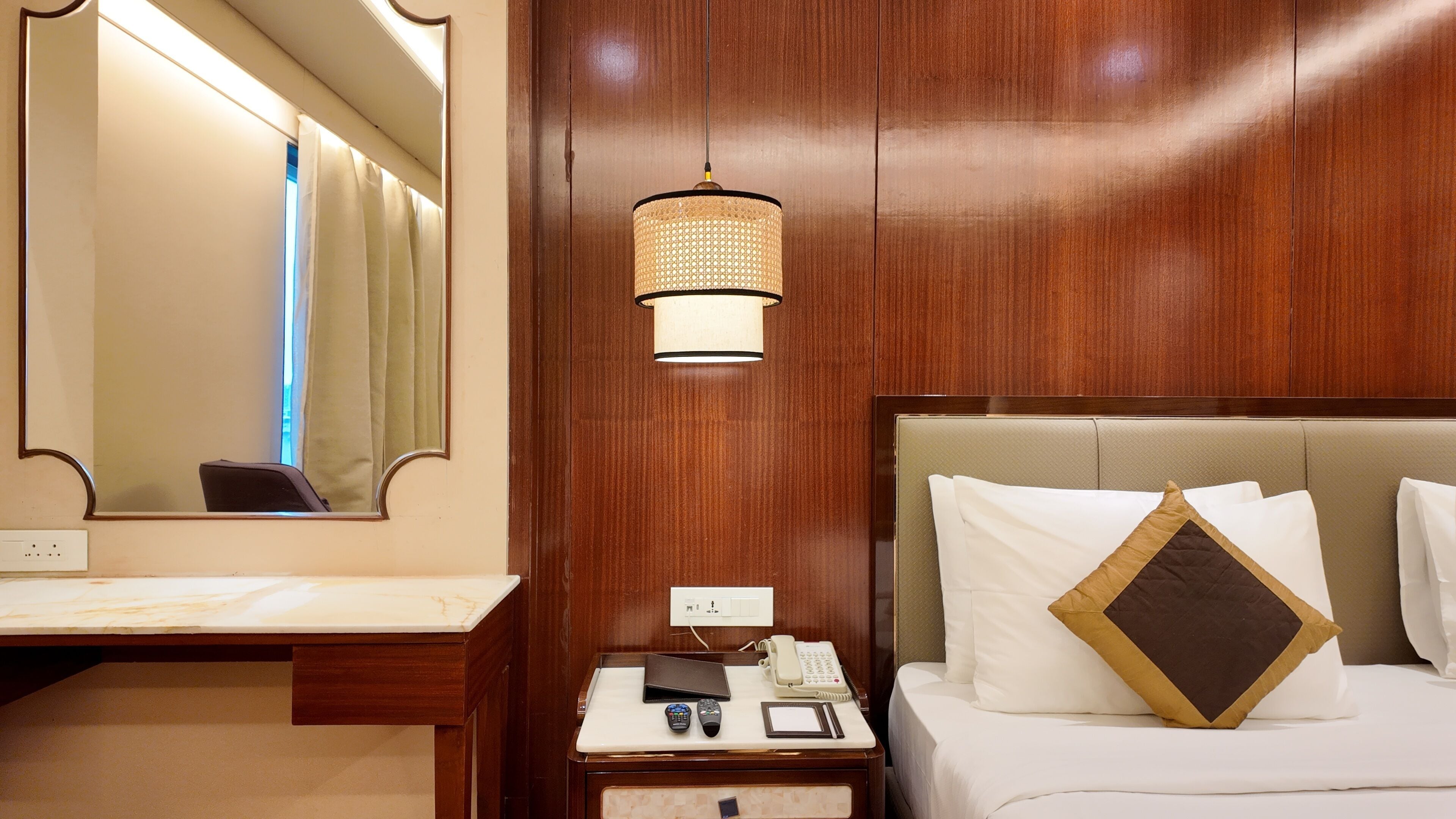 Habitación de hotel con pared de paneles de madera, un espejo grande, una mesita de noche con teléfono, control remoto y una lámpara de noche.