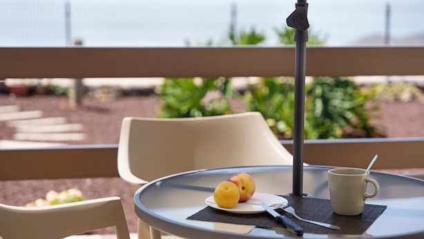 Mesa redonda con un plato de fruta, una taza y cubiertos en un balcón con vistas a un jardín.