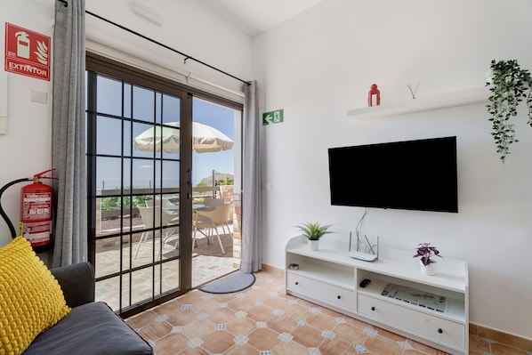 A hotel room with a sliding glass door to an outdoor patio, a flat-screen TV mounted on the wall, a white console table with a plant, and a fire extinguisher on the wall.