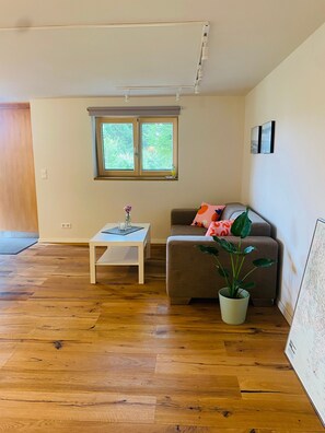 Una habitación con piso de madera, una mesa blanca, un sofá marrón, una ventana y una planta en maceta.