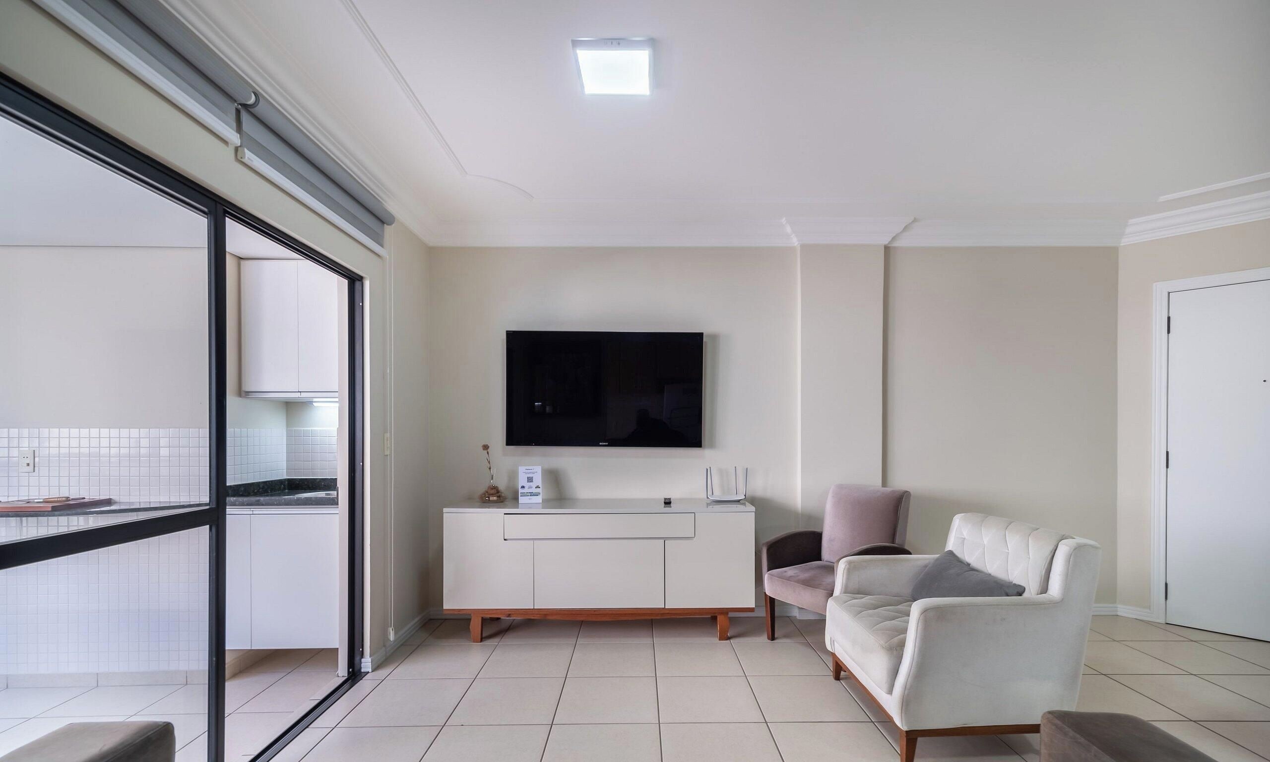 Uma sala de estar moderna com um sofá branco, uma televisão de tela plana na parede e uma porta de vidro deslizante que dá para uma varanda.