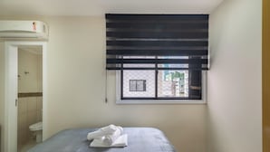 A hotel room with a bed, a window with a black blind, a wall-mounted air conditioner, and a view of buildings outside. - Sea View Apt, Priv. Loc. 2min fr th sea BC Esa0902 (Balneário Camboriú)