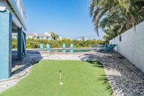 Un espace piscine comprenant un green d’entraînement au golf, des chaises longues et un palmier.
