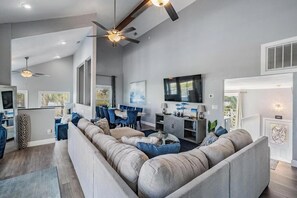 Un salon moderne comprenant un canapé d’angle, un coin repas, des ventilateurs de plafond et un téléviseur.