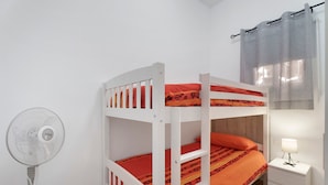 Una cama litera con ropa de cama naranja, una mesita de noche blanca con una lámpara y un ventilador de pie.