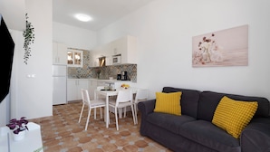 Una cocina moderna con armarios blancos, una zona de comedor con sillas blancas, un sofá gris con cojines amarillos y un cuadro colgado en la pared.