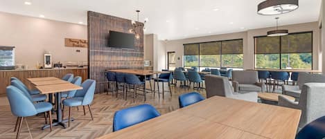 A modern dining area with a wooden table and blue chairs, a bar area with stools, and large windows offering a view of greenery.