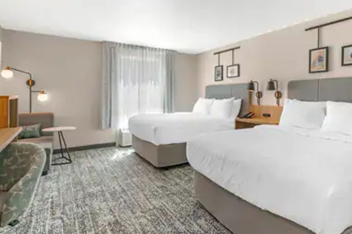 A hotel room with two beds, a desk, a chair, a lamp, and a window with curtains.