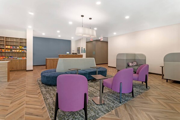 A modern lounge area with a blue sofa, purple chairs, a small table, and a wooden bookshelf with snacks. - Sleep Inn & Suites (Ashland City)