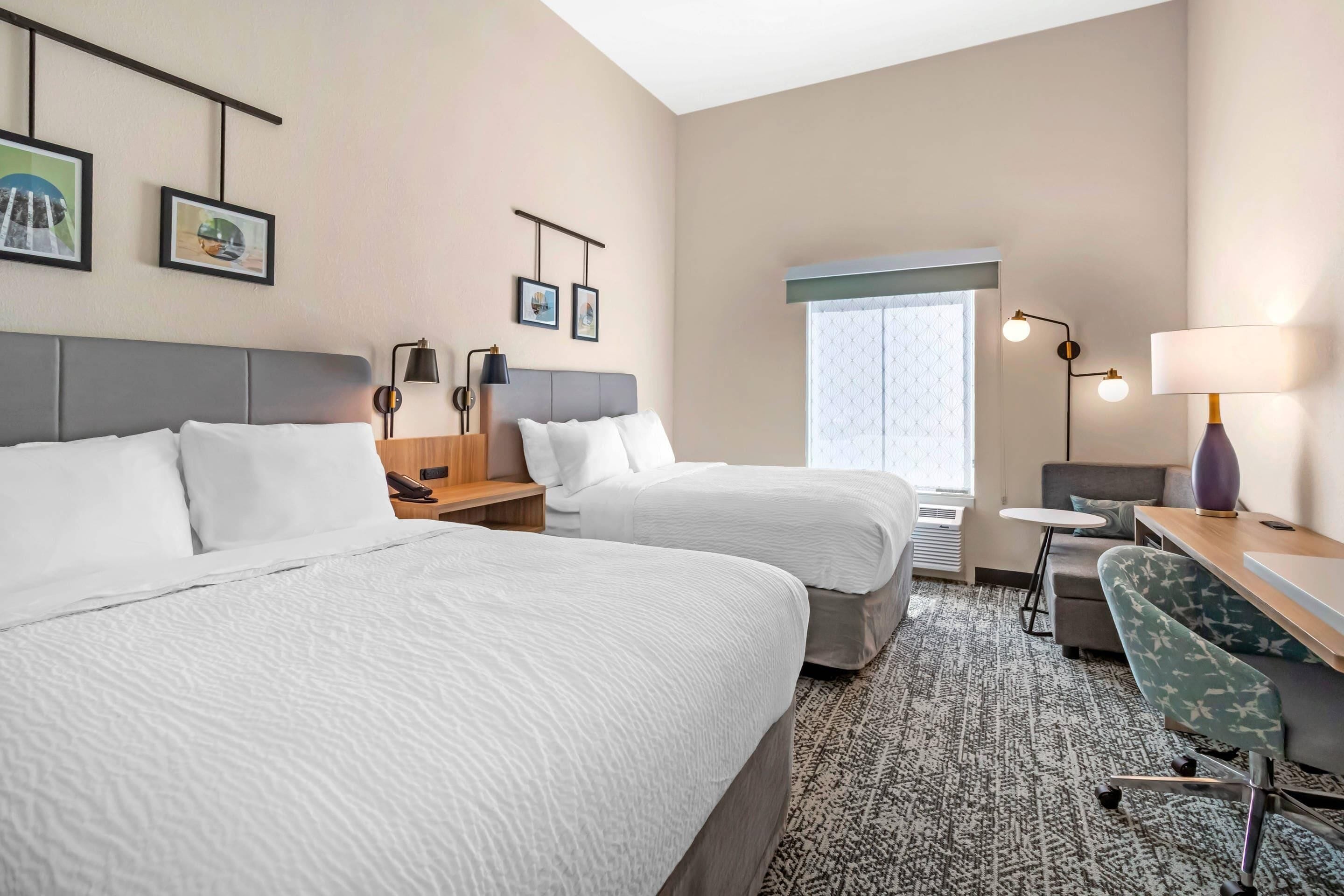 A hotel room with two beds, a desk, a chair, a lamp, and framed pictures on the wall.