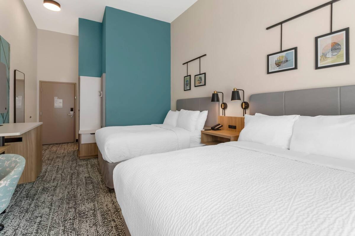 Une chambre d’hôtel avec deux lits, un bureau et une chaise. Les murs sont ornés de tableaux encadrés et la porte est munie d’une pancarte.