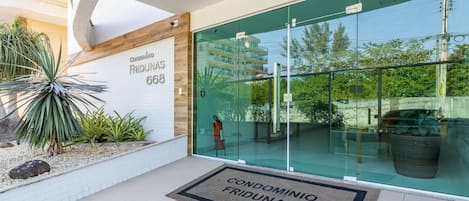 A modern building entrance with a glass door, a doormat with the building's name, and a small garden area with plants.
