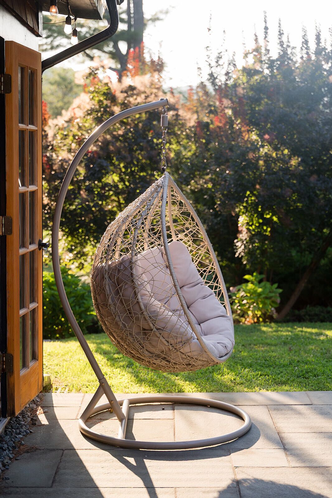 Una silla colgante con asiento acolchado y estructura metálica, ubicada en un patio con puerta de madera y jardín al fondo.