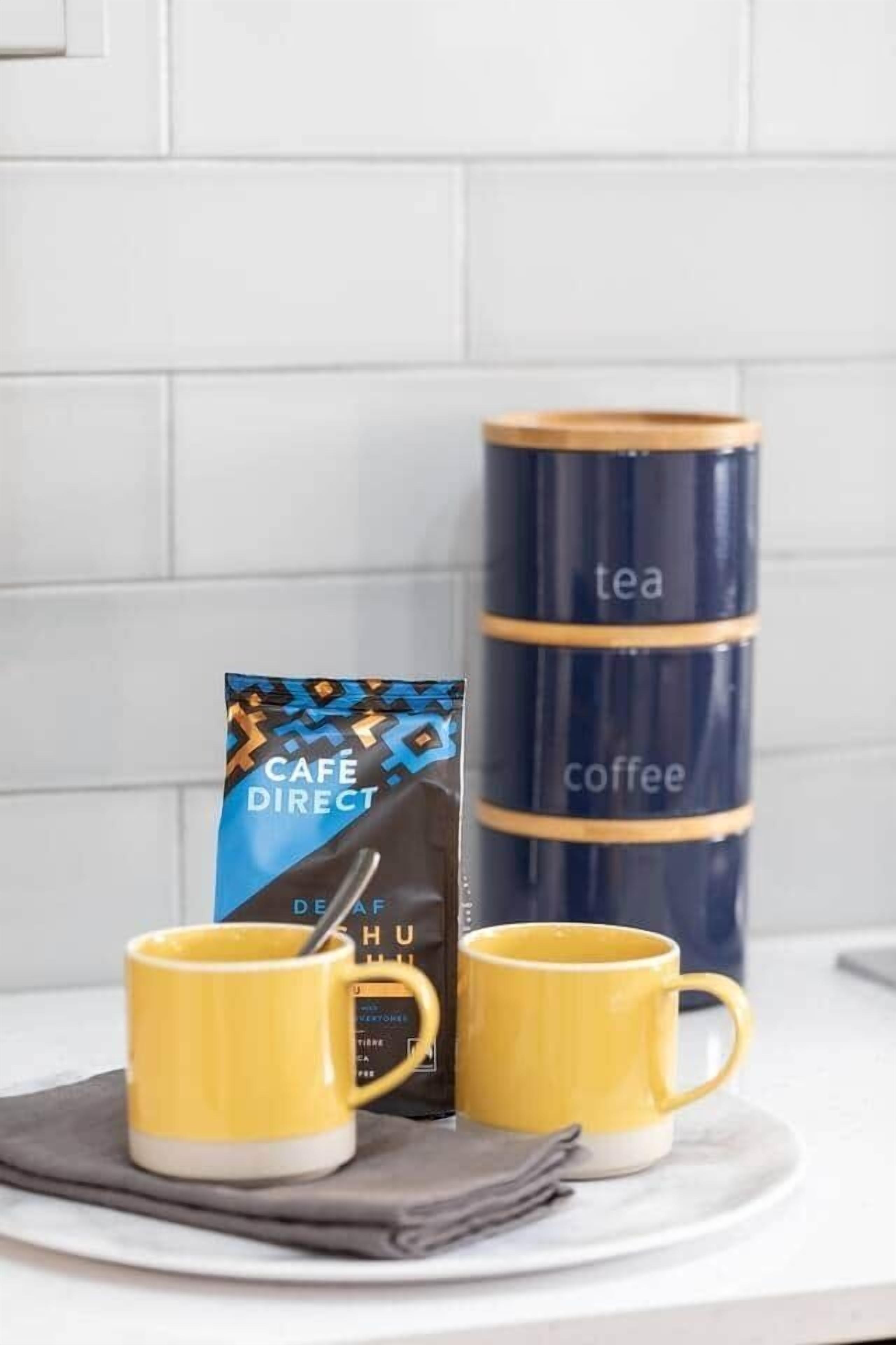 Twee gele mokken op een wit bord, een blauwe en zwarte koffiecontainer, en een koffieverpakking met een geometrisch patroon.