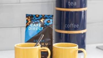 Twee gele mokken op een wit bord, een blauwe en zwarte koffiecontainer, en een koffieverpakking met een geometrisch patroon.