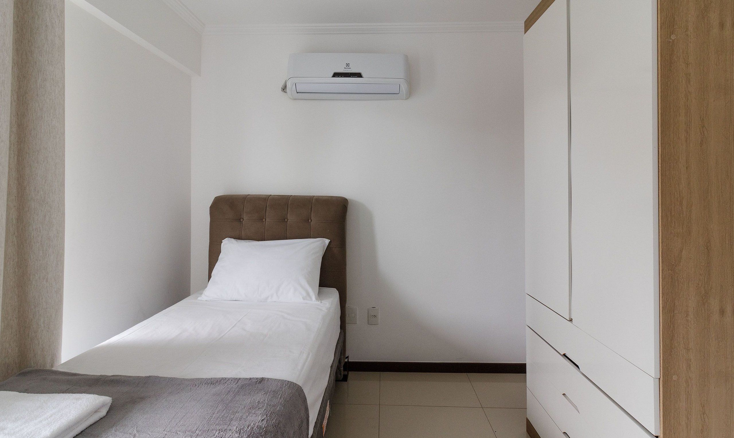 Una habitación pequeña y ordenada con una cama individual, aire acondicionado de pared y un panel de madera en una pared.