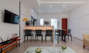 A modern kitchen with a central island, a dining area with four chairs, a TV mounted on a white brick wall, and a wooden bench with a ship model. - Apt 400m From the Beach of Taperapua/ BA Edt0011 (Porto Seguro)