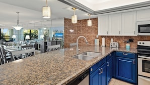 A modern kitchen with granite countertops, a stainless steel sink, blue cabinets, and a dining area with a glass-top table and chairs. - Steps to Pool & Beach: South Padre Island Retreat! (South Padre Island)