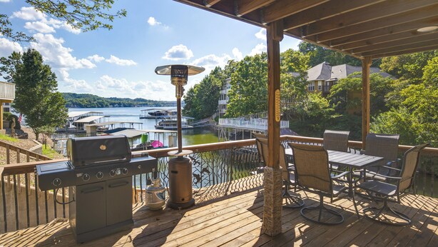 A wooden deck with a grill, table, and chairs overlooking a lake with boats and houses in the background. - Fire Pit + Private Dock: Ozarks Family Escape! (Camdenton)