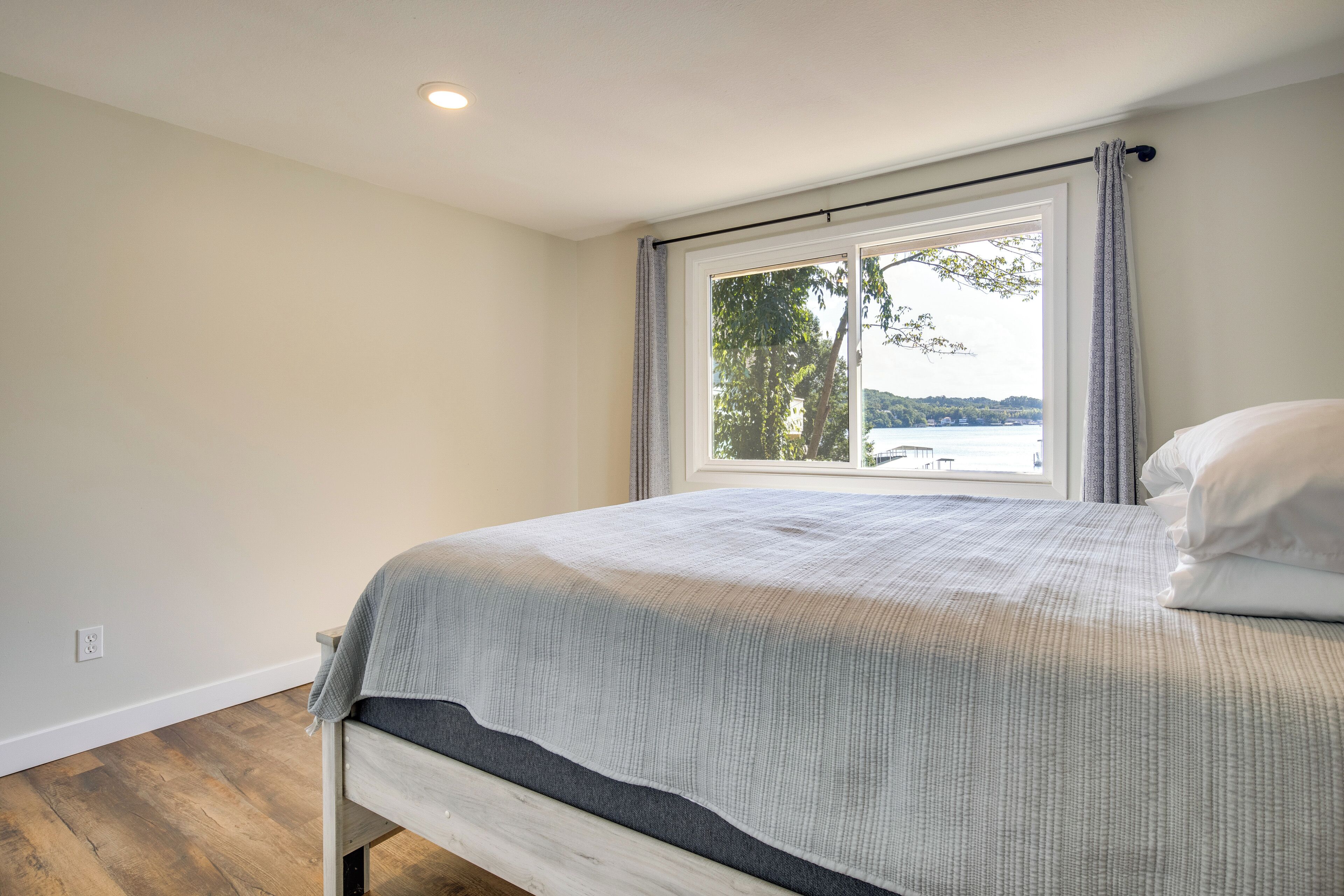 Una camera da letto con un'ampia finestra, un letto con copripiumino grigio e vista su un corpo d'acqua e alberi.