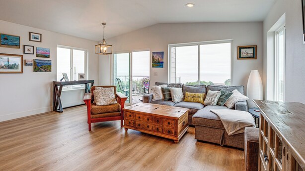 A spacious living room with a wooden coffee table, a sofa, and a chair. There are large windows providing natural light and a view of the outdoors.