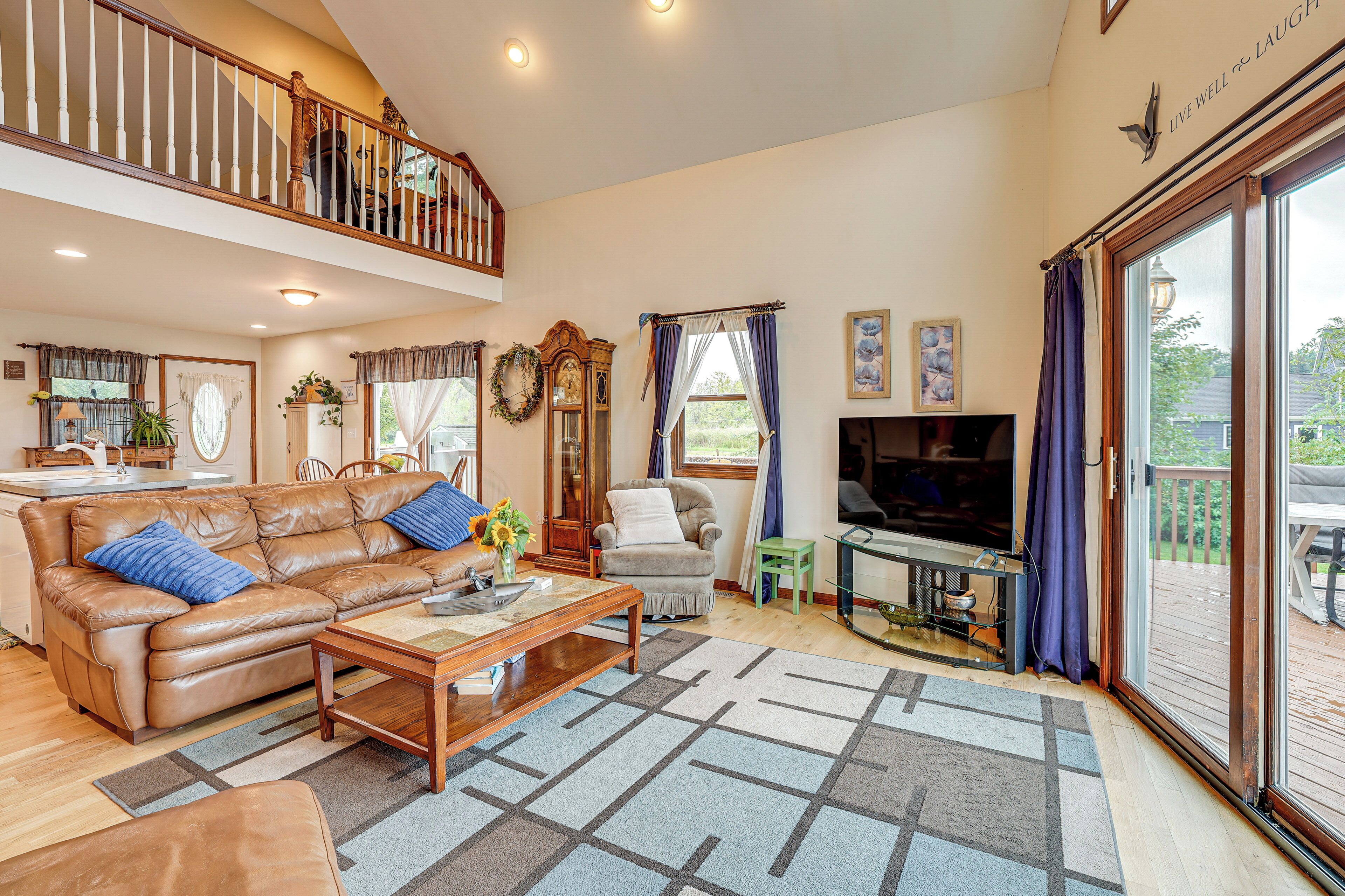 A spacious living room with a brown leather sofa, a wooden coffee table, and a television. There is a staircase leading to an upper level, and a large window with curtains.
