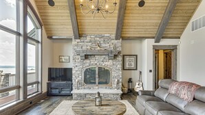 A spacious living room with a stone fireplace, leather sofa, and large windows overlooking a scenic view. - River Views + Wraparound Deck: Dubuque Getaway (Dubuque)