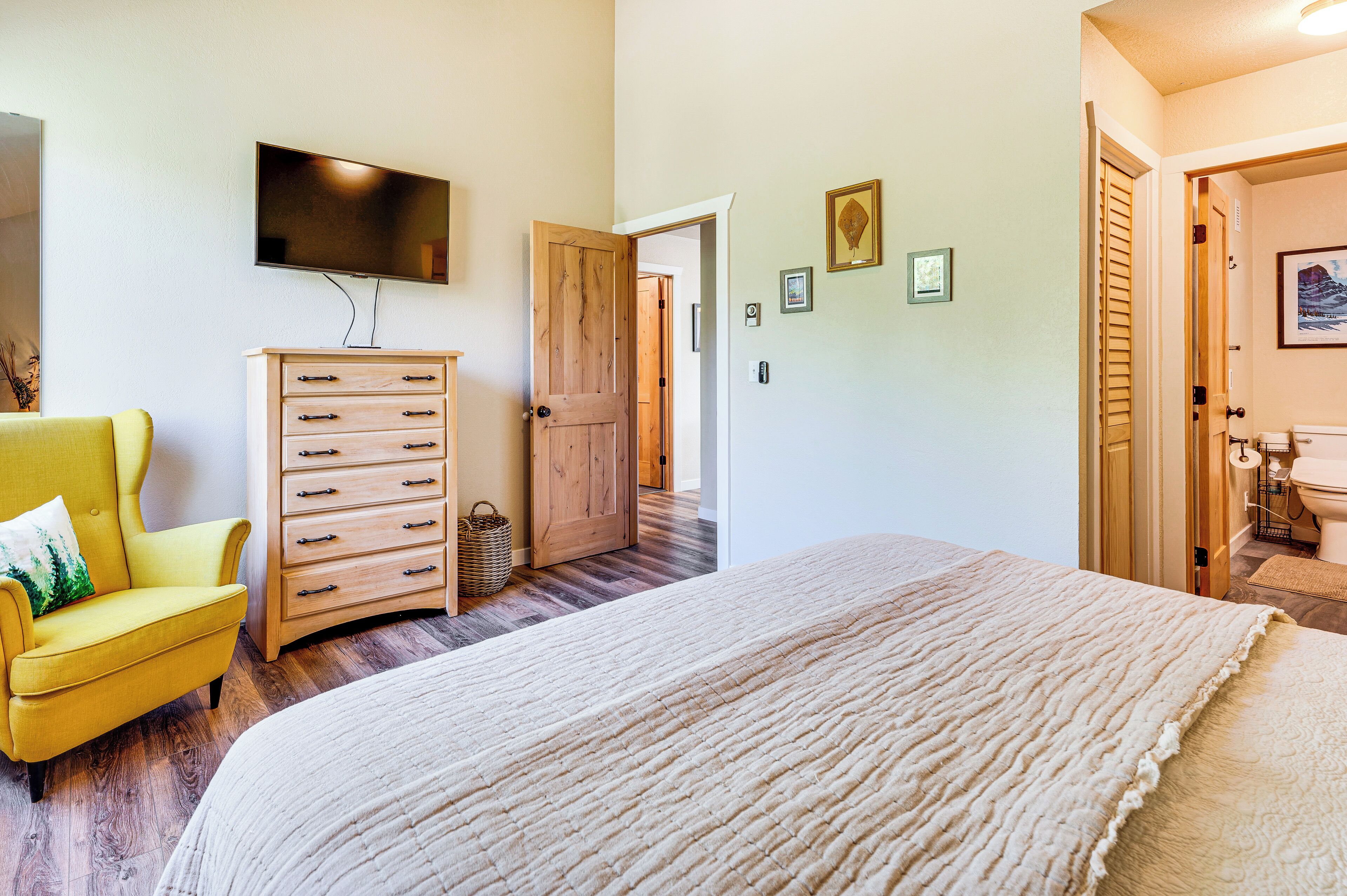 A bedroom with a bed, a wooden dresser, a yellow armchair, a television on the wall, and a door leading to another room.