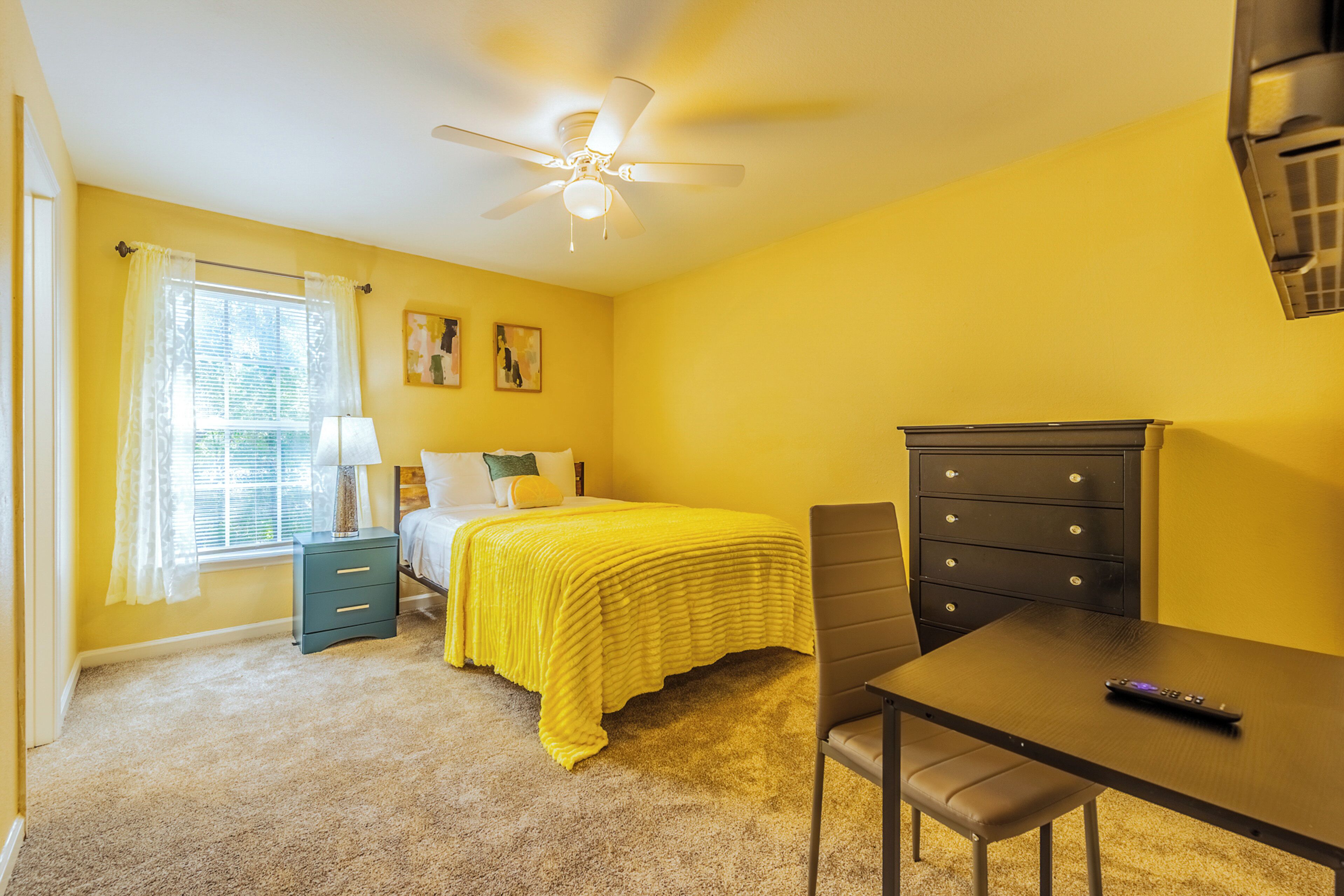A bedroom with a bed, a desk, a chair, a dresser, a ceiling fan, and a window with curtains.