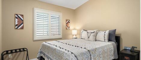 A bedroom with a bed, a nightstand, a lamp, a clock, a window with blinds, and a ceiling fan.