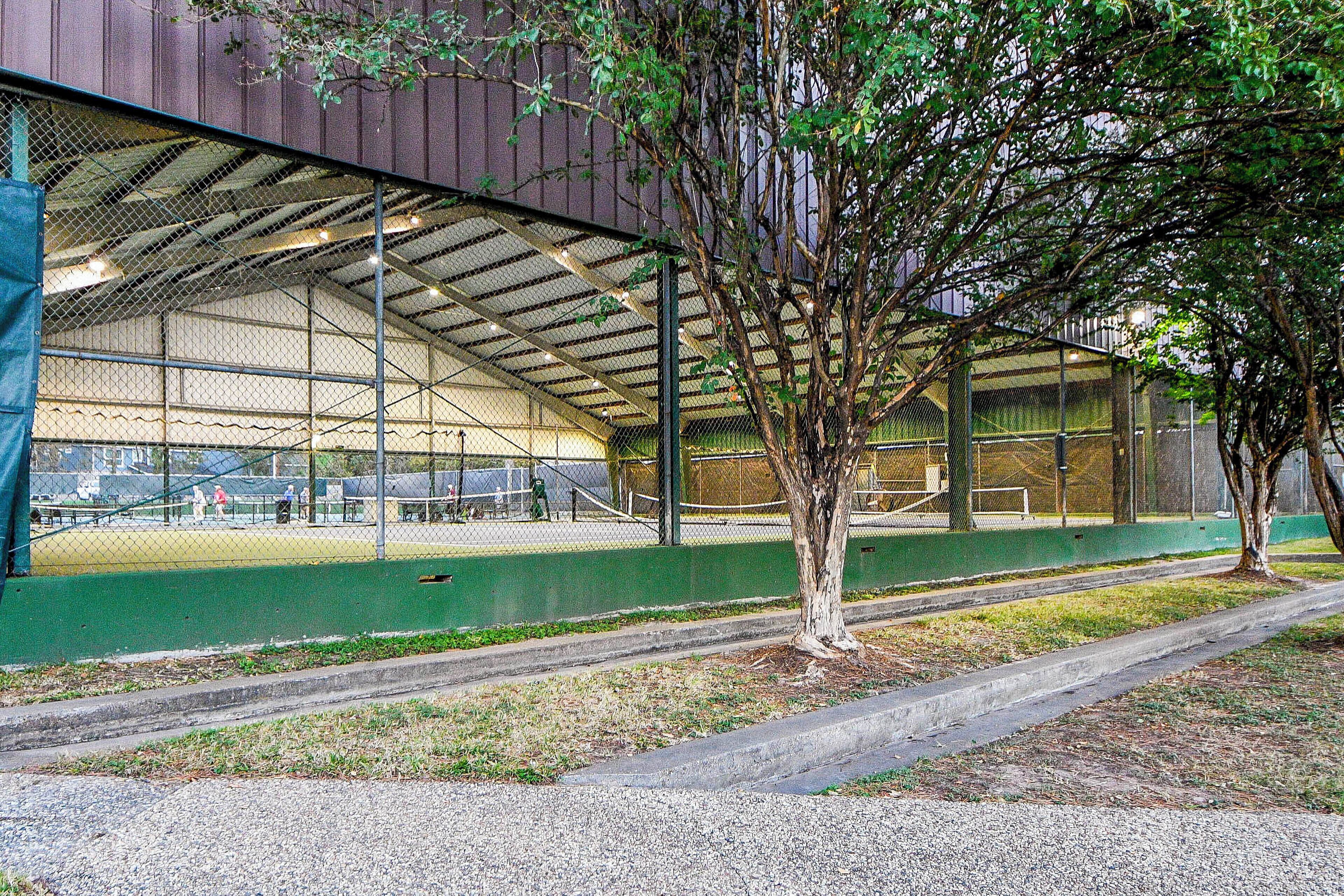A covered sports court with a netted roof, surrounded by trees and a gravel path.