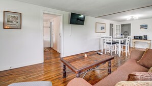 Un salon comprenant une table basse en bois, un canapé, un coin repas avec une table et des chaises blanches, et un téléviseur à écran plat fixé au mur.